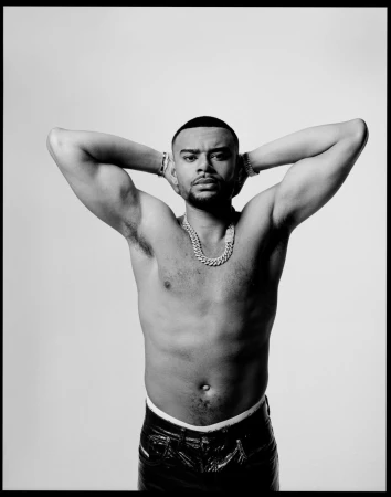 Black and white portrait of a shirtless man with arms raised, wearing a thick chain necklace and dark trousers against a light background.