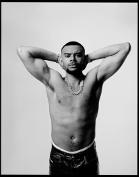 Black and white portrait of a shirtless man with arms raised, wearing a thick chain necklace and dark trousers against a light background.