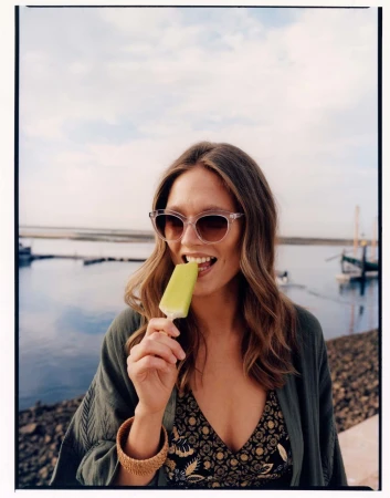 Woman wearing sunglasses and a green cardigan eating a yellow popsicle by the water