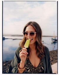 Woman wearing sunglasses and a green cardigan eating a yellow popsicle by the water