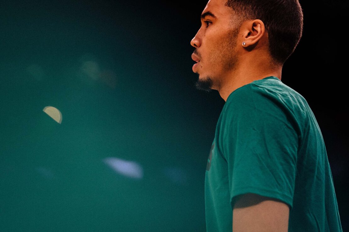 Profile shot of an adult male with dark hair and a beard, wearing a green t-shirt, against a dark green and black bokeh background.