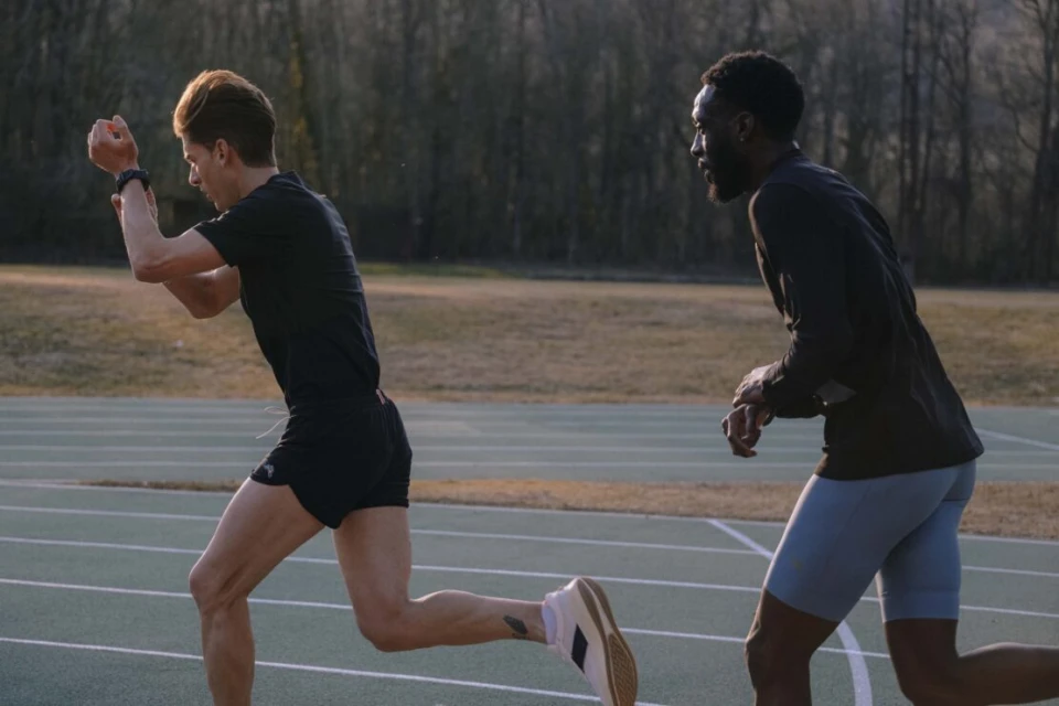 Two male athletes, one light-skinned and one dark-skinned, run on a green track during the daytime, with a blurred tree background.
