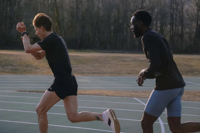 Two male athletes, one light-skinned and one dark-skinned, run on a green track during the daytime, with a blurred tree background.