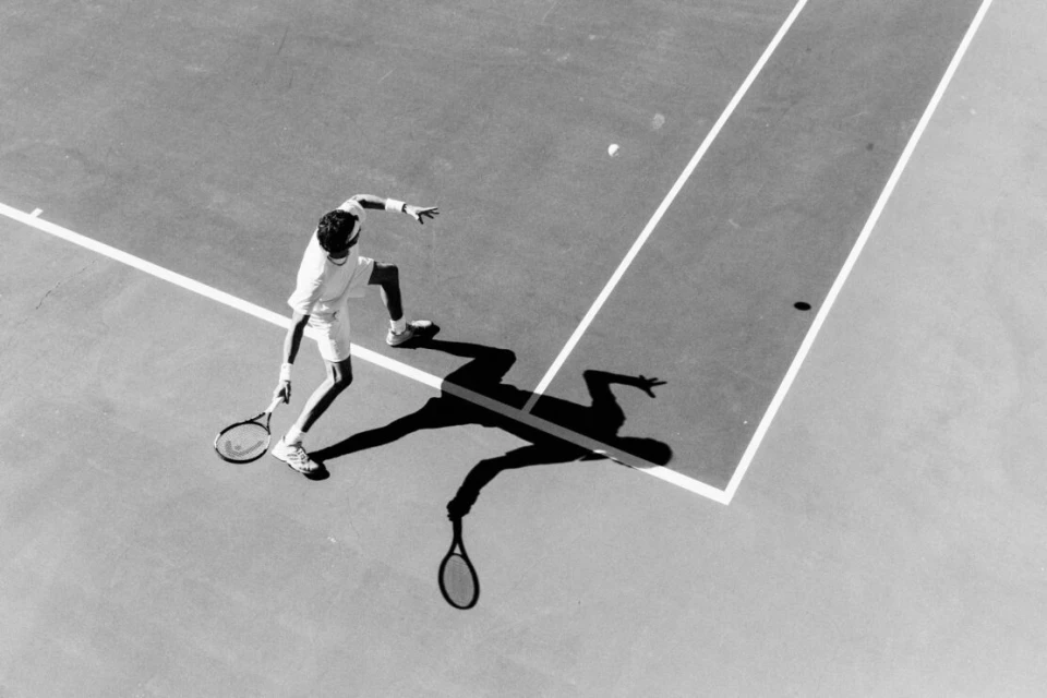Monochrome aerial view of an adult tennis player serving a ball on a court, showcasing action, shadow, and court lines.