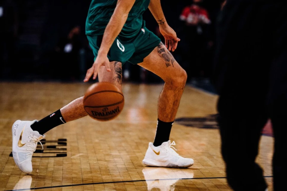 A basketball player in a green uniform dribbles a basketball on a hardwood court, wearing white shoes with black socks.