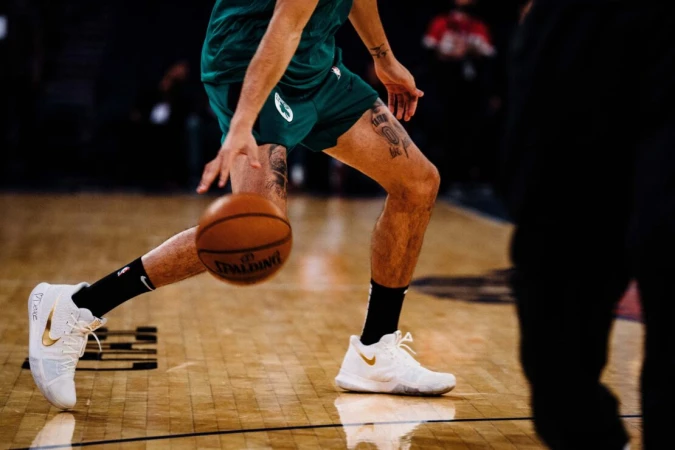 A basketball player in a green uniform dribbles a basketball on a hardwood court, wearing white shoes with black socks.