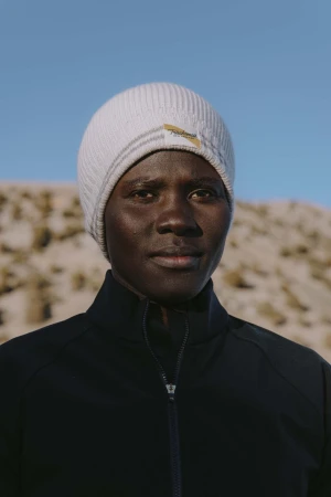Portrait of a dark-skinned adult male in a black jacket and white beanie against a blurred, natural outdoor background and clear blue sky.
