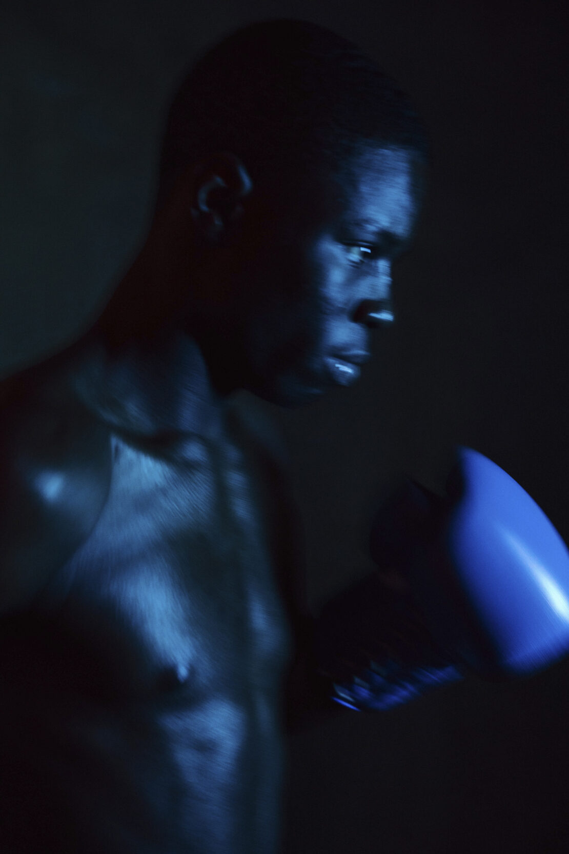Low-key portrait of an African American male boxer with blue boxing glove, partially shirtless, under blue-toned lighting in a dark environment.