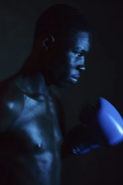 Low-key portrait of an African American male boxer with blue boxing glove, partially shirtless, under blue-toned lighting in a dark environment.