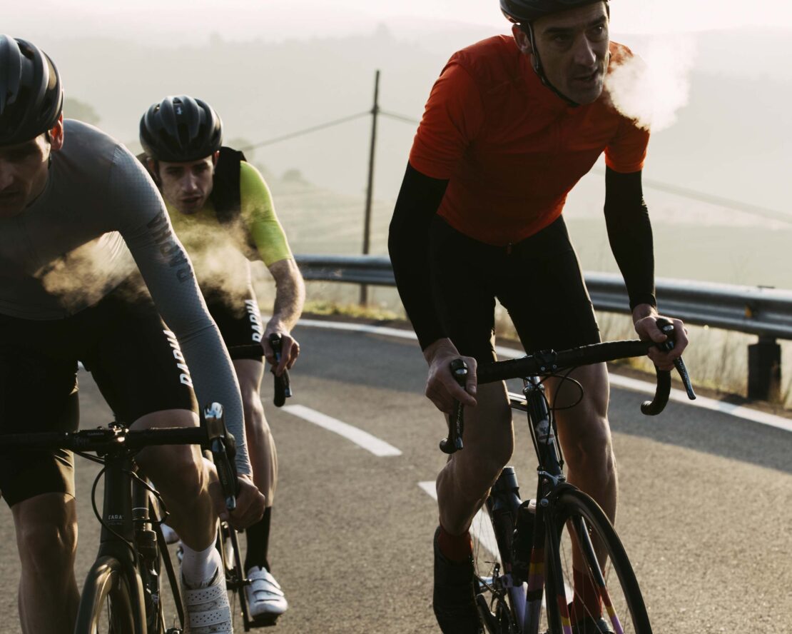 Three cyclists ride bicycles uphill on a paved road with rolling hills in the background, showcasing a challenging outdoor sport.