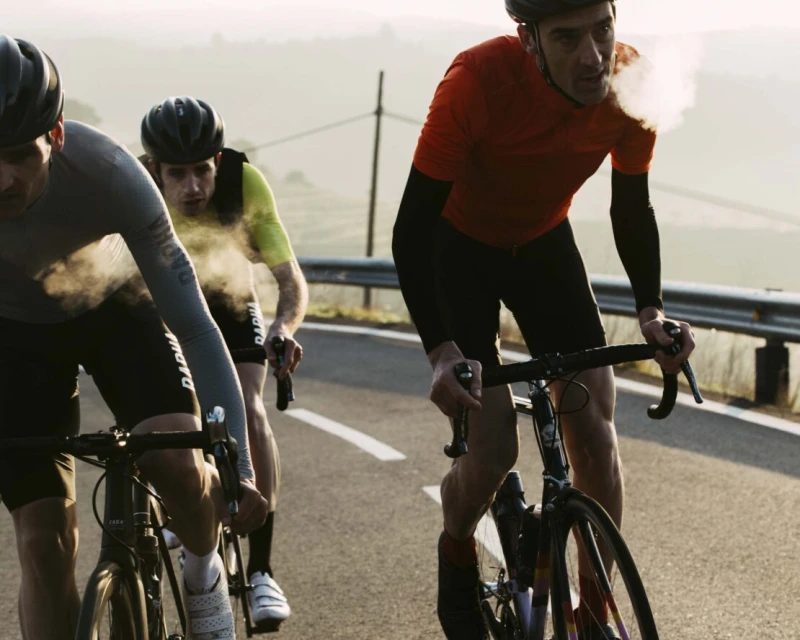 Three cyclists ride bicycles uphill on a paved road with rolling hills in the background, showcasing a challenging outdoor sport.