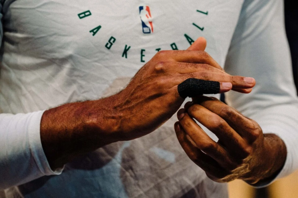 Close-up shot of a basketball player wrapping tape around a finger, wearing a white long-sleeved shirt with 'BASKETBALL' printed on it.