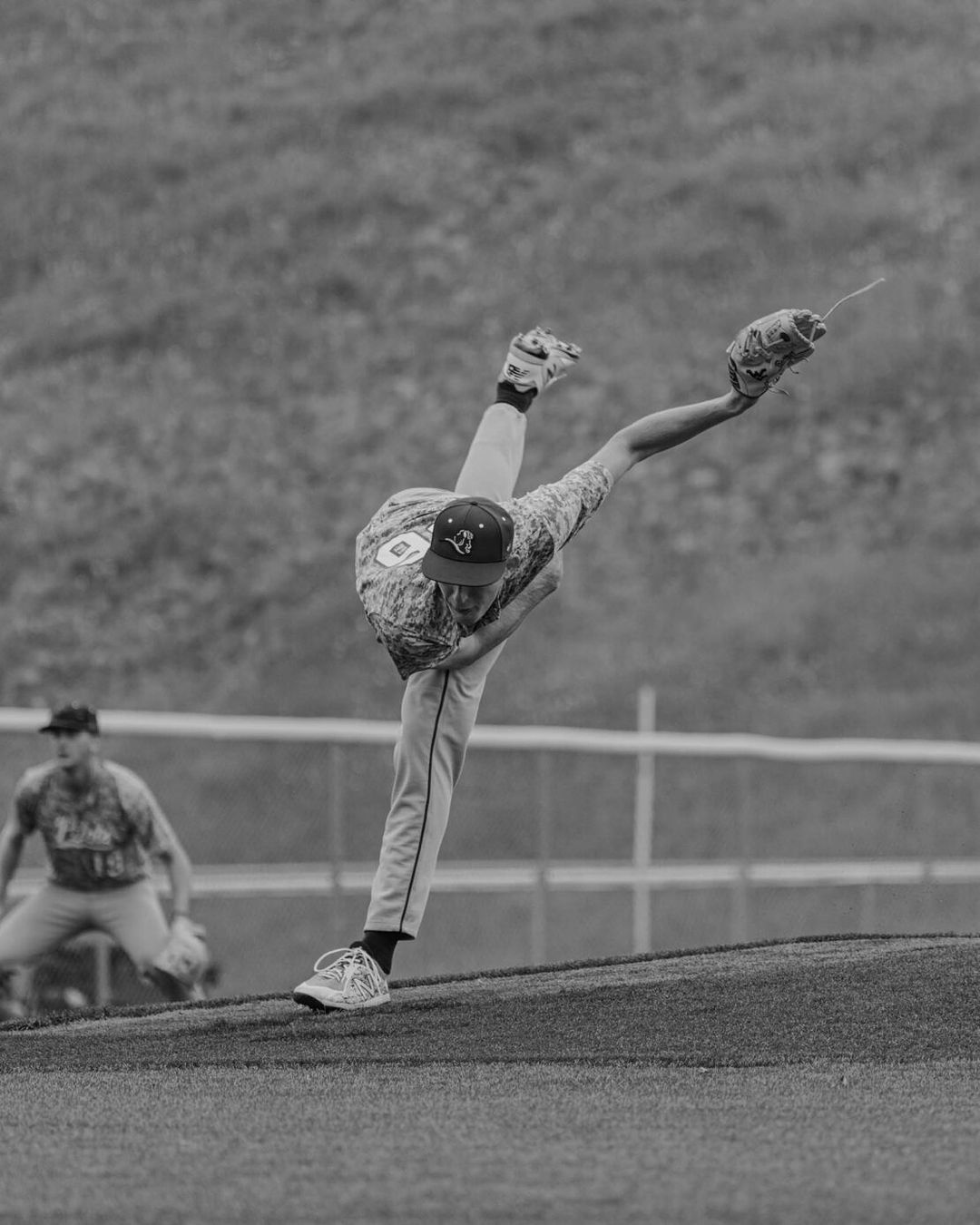 Monochrome action shot of a baseball pitcher throwing a ball on a grassy field with another player and grassy hillside in the background.
