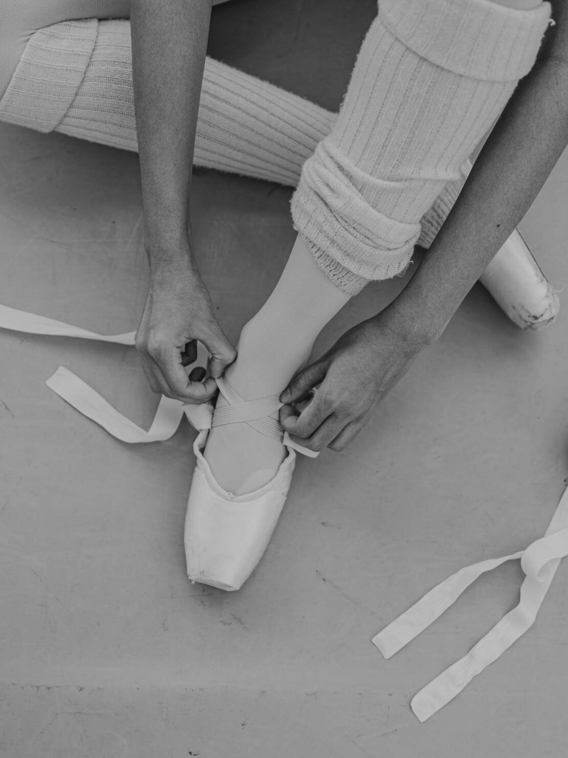 Close-up of a dancer tying ribbons on pointe shoes wearing ribbed leg warmers in a monochrome photograph by Emily Maye.
