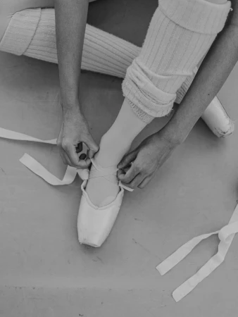 Close-up of a dancer tying ribbons on pointe shoes wearing ribbed leg warmers in a monochrome photograph by Emily Maye.