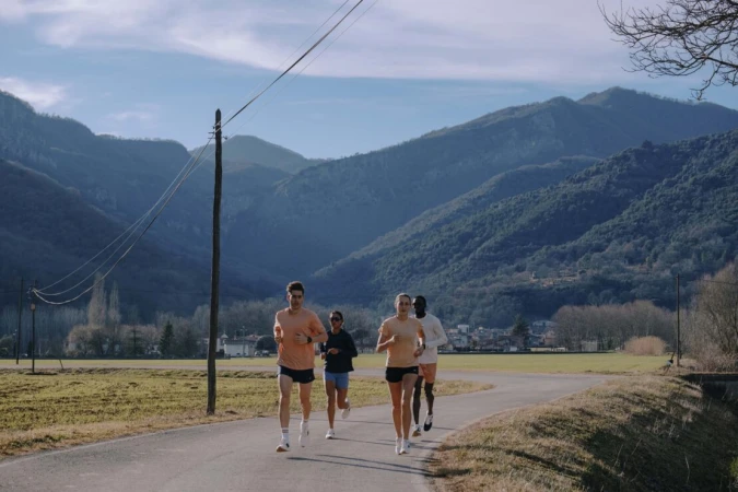 Group of four athletes running on paved road with green fields and mountain range backdrop under sunny sky. Outdoor fitness scene.