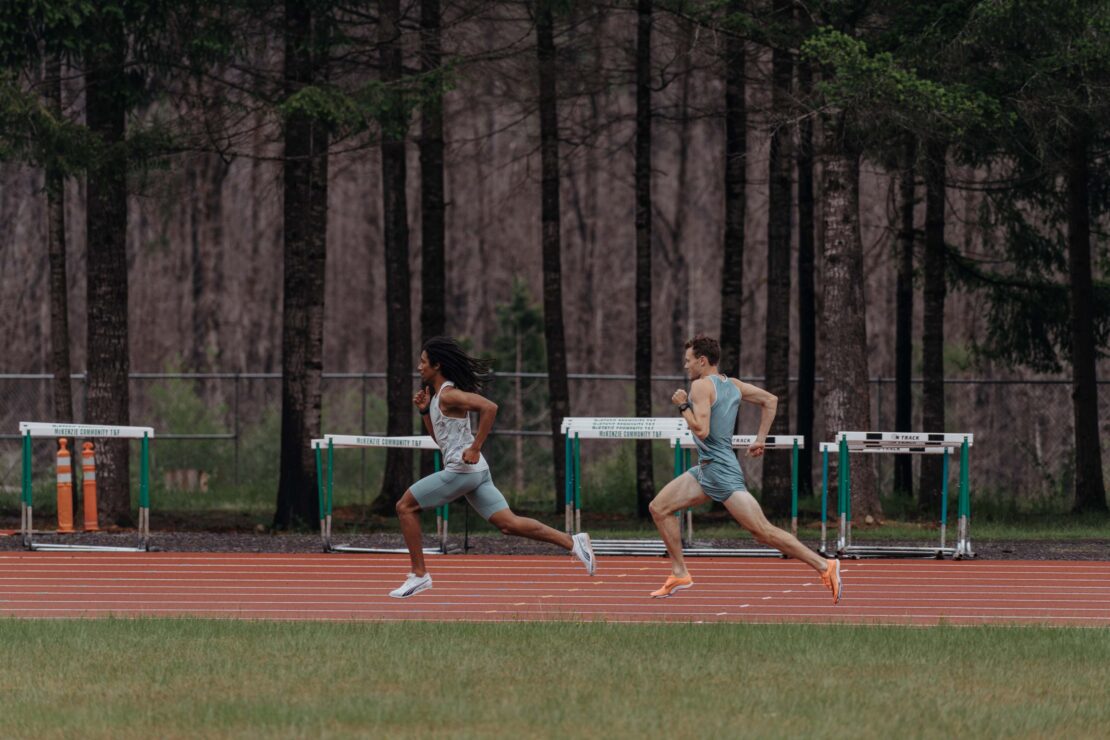 Two athletic men sprinting on a red track in a verdant outdoor setting with a backdrop of trees and hurdles, showcasing fitness and speed.