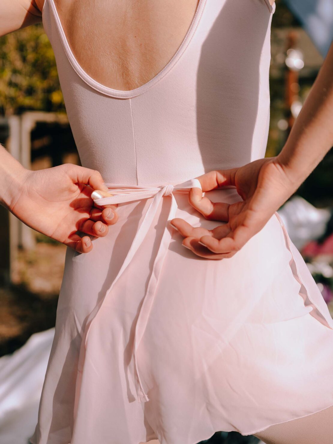Back view of a woman in a pink leotard and skirt tying a sash in an outdoor setting. Light and airy fashion and dance attire.