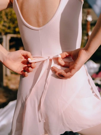 Back view of a woman in a pink leotard and skirt tying a sash in an outdoor setting. Light and airy fashion and dance attire.