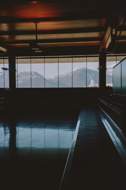 Interior view of an empty building with large windows overlooking snow-capped mountains, reflecting on a polished floor, under wood ceiling.