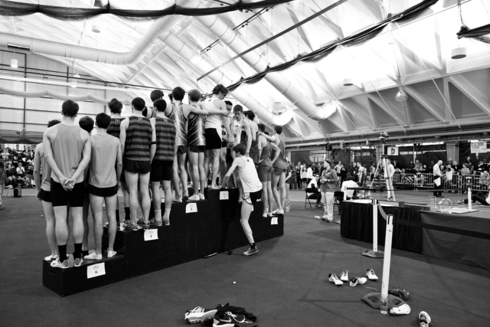 A group of young male athletes stand on a numbered podium in an indoor stadium with track, with spectators in background.
