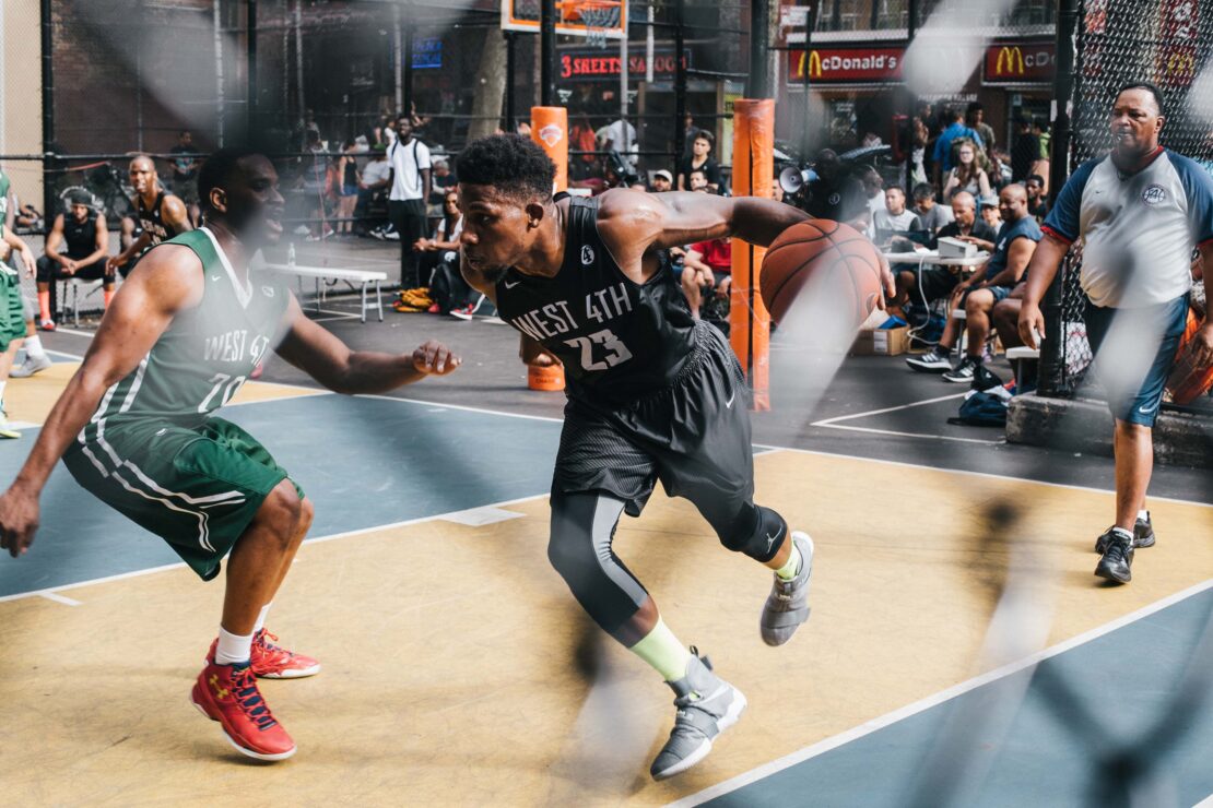 An outdoor action shot of two basketball players in uniforms playing on a court, with spectators and McDonald's in the background.
