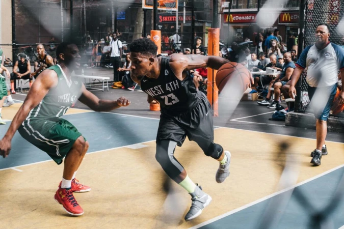 An outdoor action shot of two basketball players in uniforms playing on a court, with spectators and McDonald's in the background.