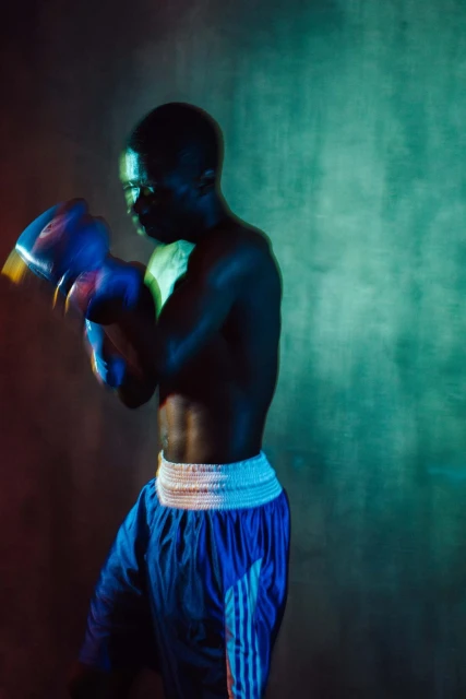 Athletic man in boxing stance with blue gloves and shorts under vibrant lighting, showcasing physical fitness and intense focus.