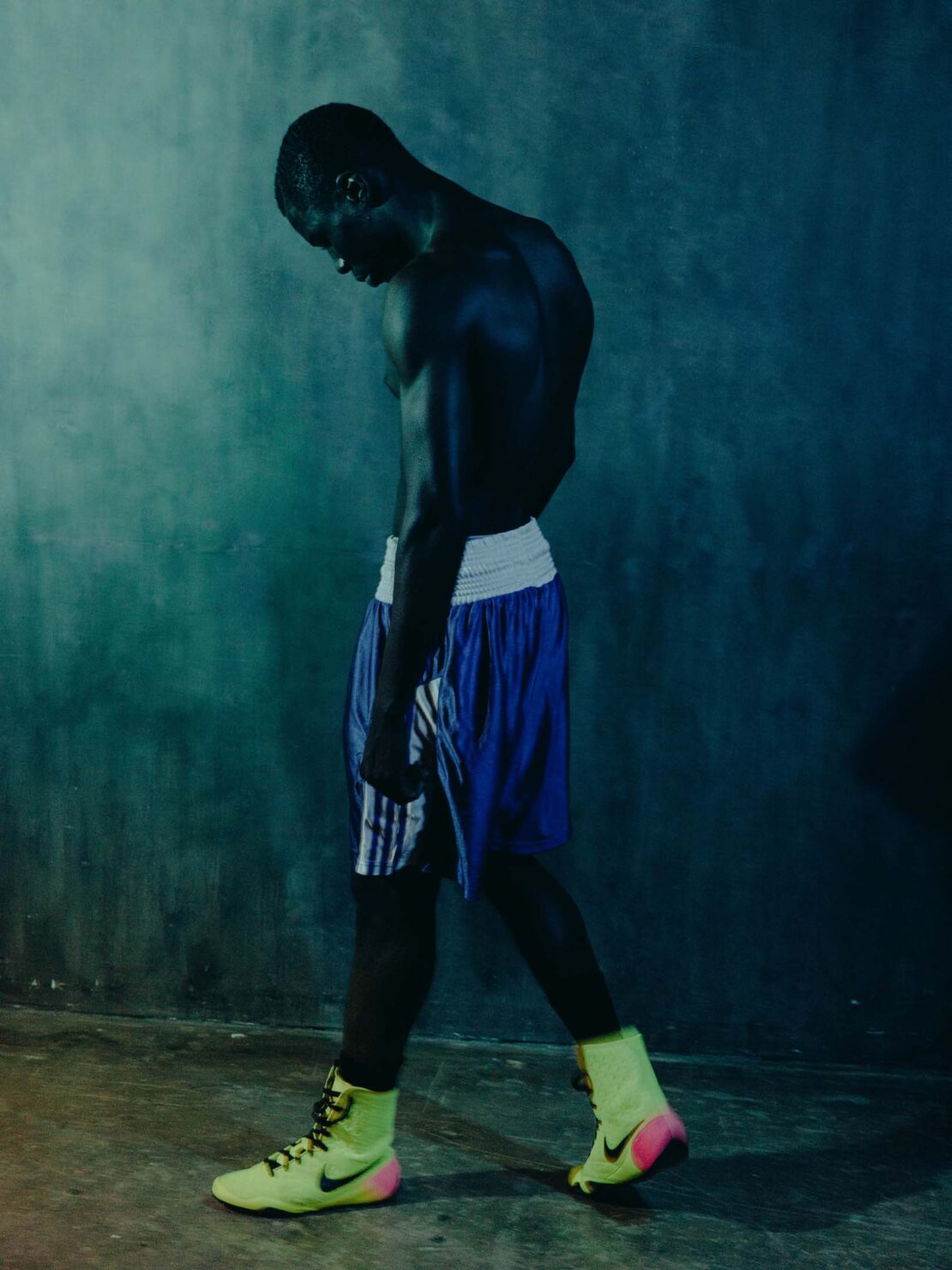Athletic Black man stands with a slight head down wearing boxing shoes and boxing shorts in front of a dark textured wall.