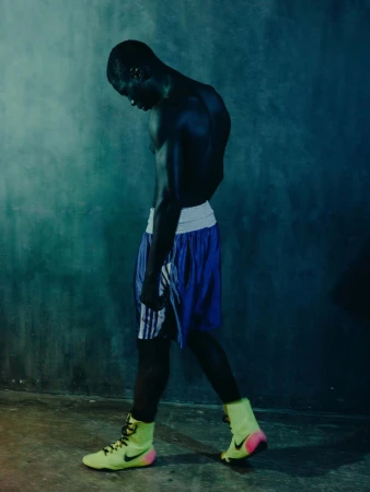 Athletic Black man stands with a slight head down wearing boxing shoes and boxing shorts in front of a dark textured wall.
