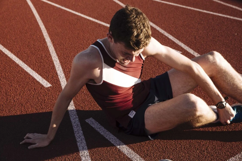 Male athlete in maroon and white tank top sits on a red running track with white lines, clutching his leg in apparent discomfort.