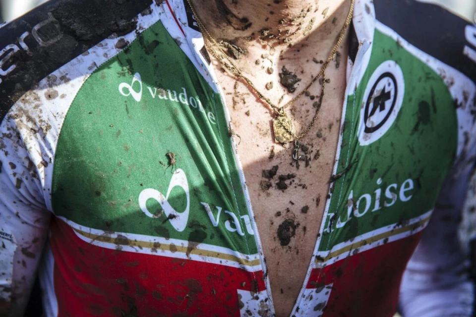 Close-up of a person's chest and neck covered in mud, wearing a green and red cycling jersey with gold necklaces.