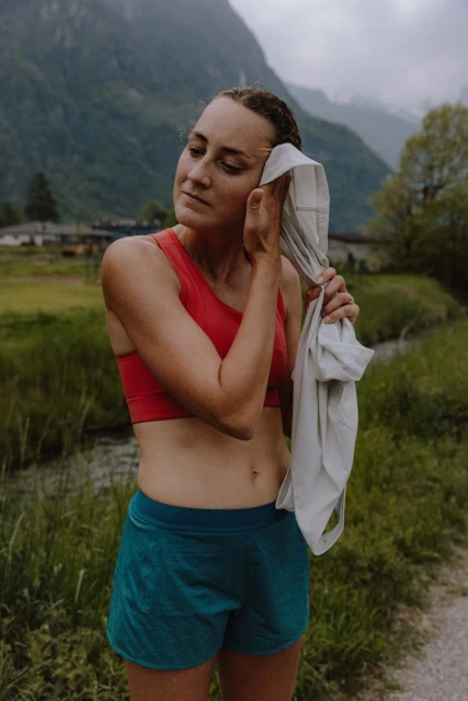 Athletic woman in sportswear wipes sweat with a towel outdoors; the background shows mountains, trees, and a small stream.
