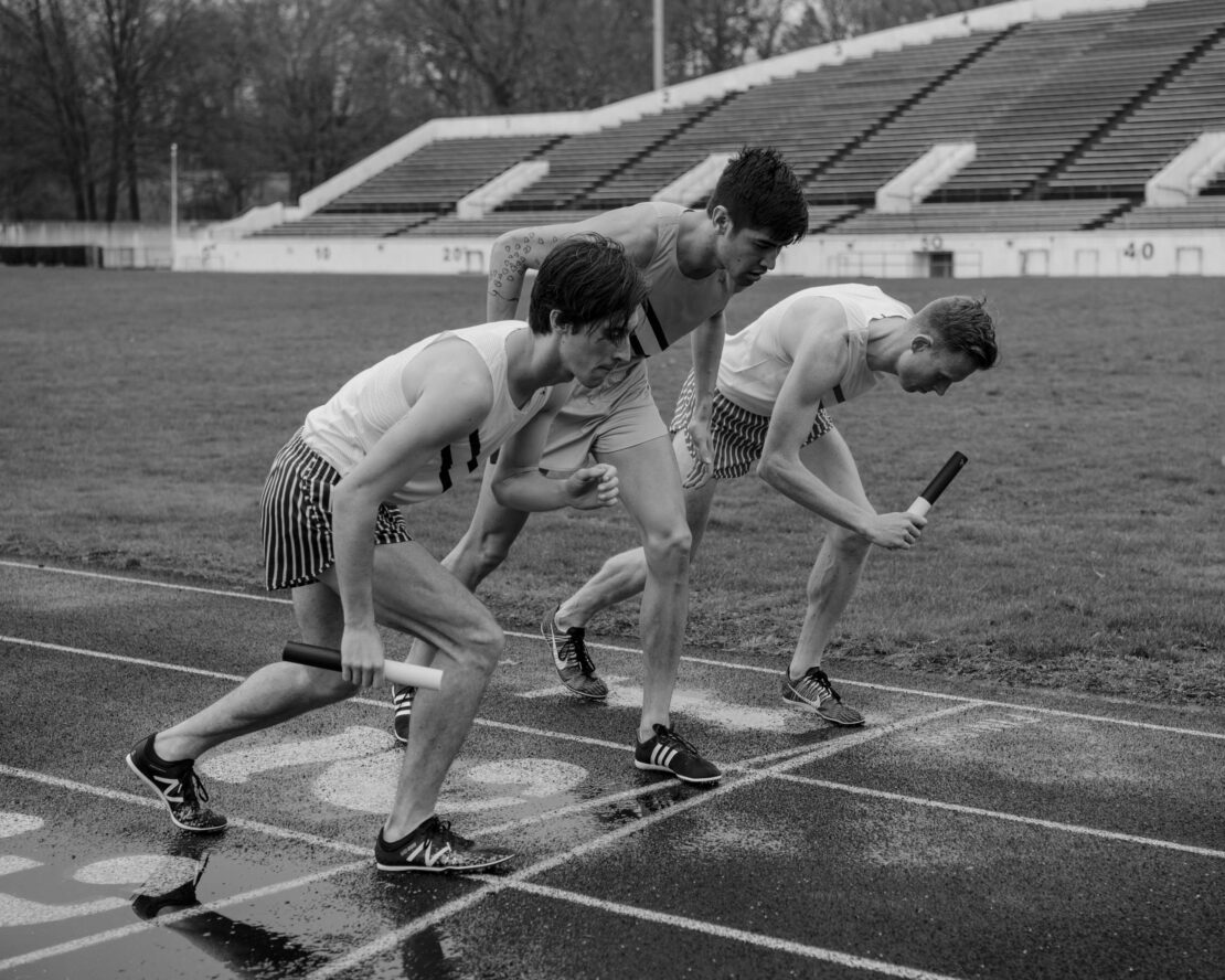 Three male athletes in track and field uniforms prepare to run a relay race outdoors on a track, stadium seating in the background.