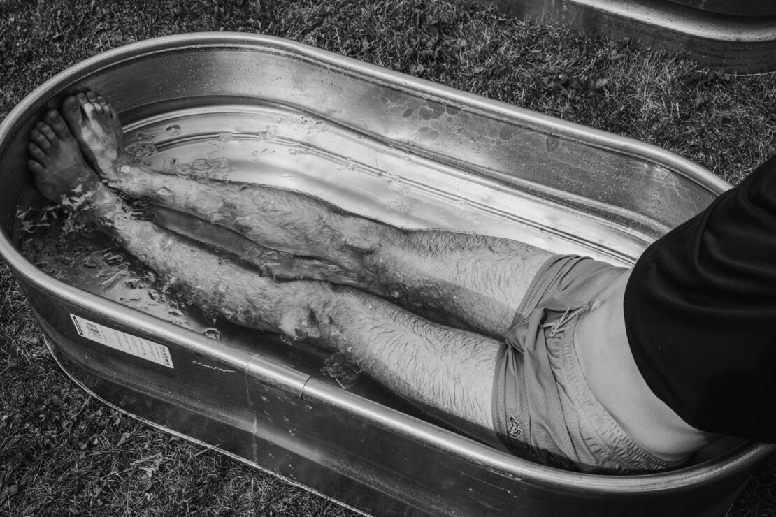 Black and white shot of a man sitting in a metal water trough outdoors, legs submerged in water; relaxation and recovery concept.