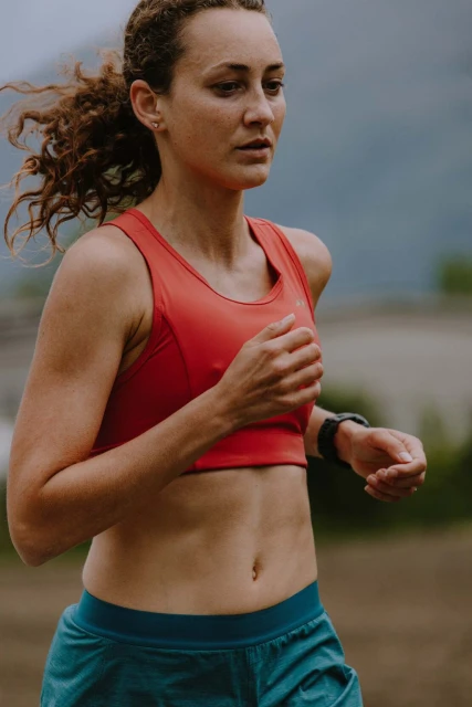 Female runner in red top and teal shorts with mountains in background, athletic wear, sportswear, fitness, exercising, running outdoors.
