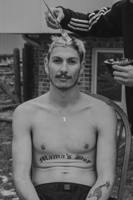 Black and white portrait of a bare-chested man sitting in a chair outdoors having his hair dyed by someone holding a brush.