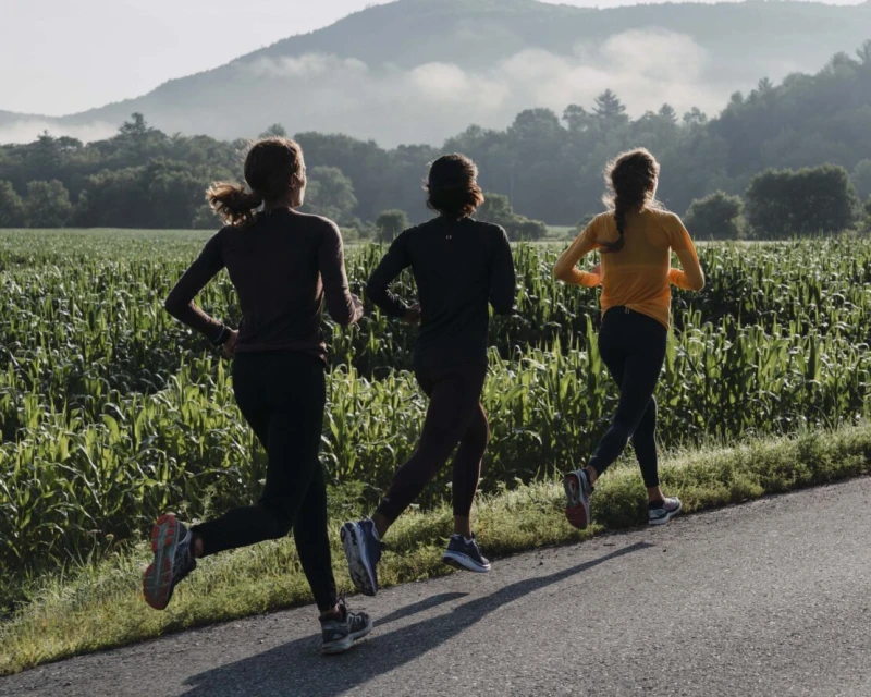 Three women jogging on an asphalt road through a rural landscape with a cornfield and distant mountains under a clear sky. Fitness lifestyle.