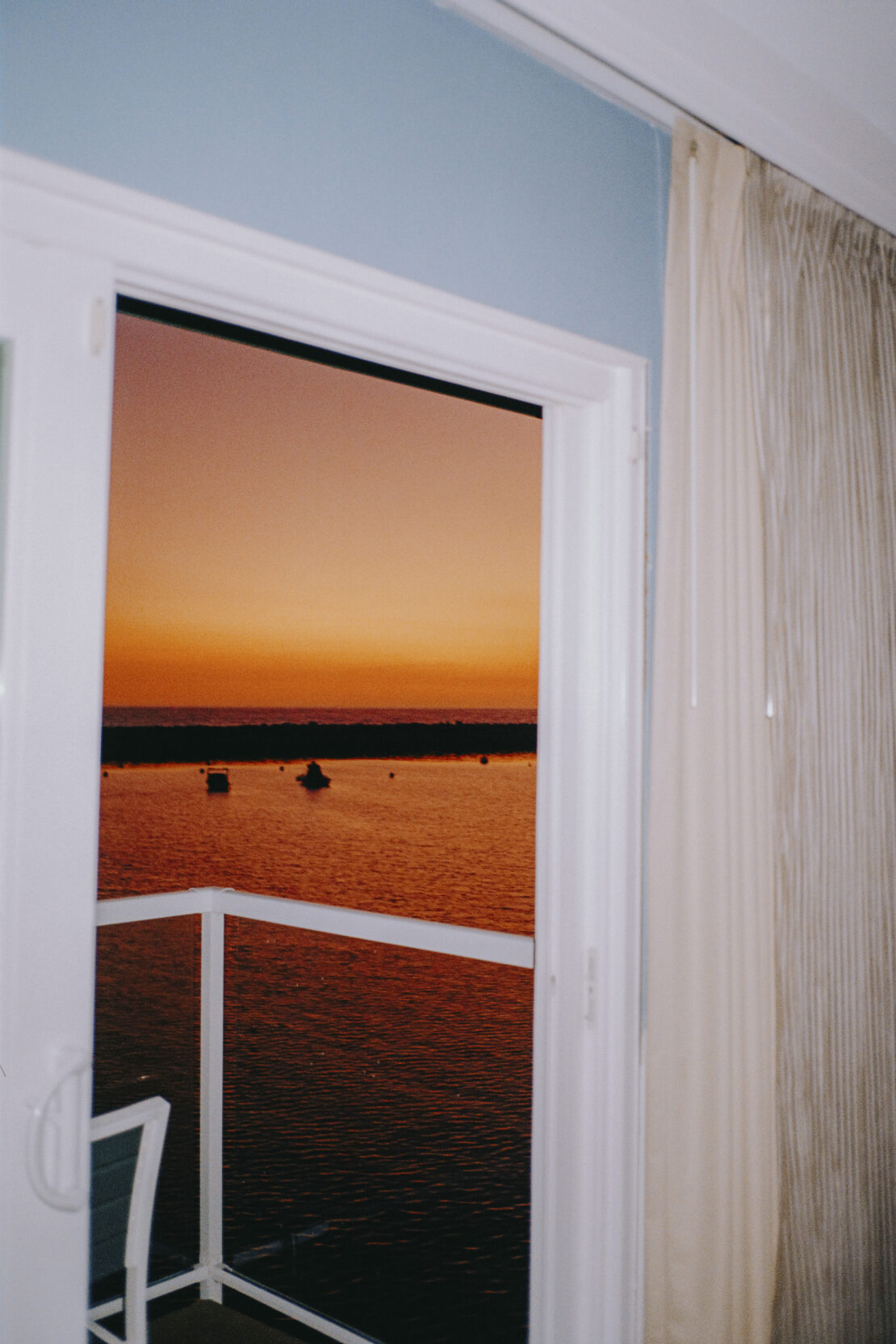 Orange sunset is visible from an open white-framed door, overlooking the water with boats, and a white curtain inside.