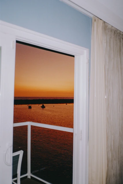 Orange sunset is visible from an open white-framed door, overlooking the water with boats, and a white curtain inside.