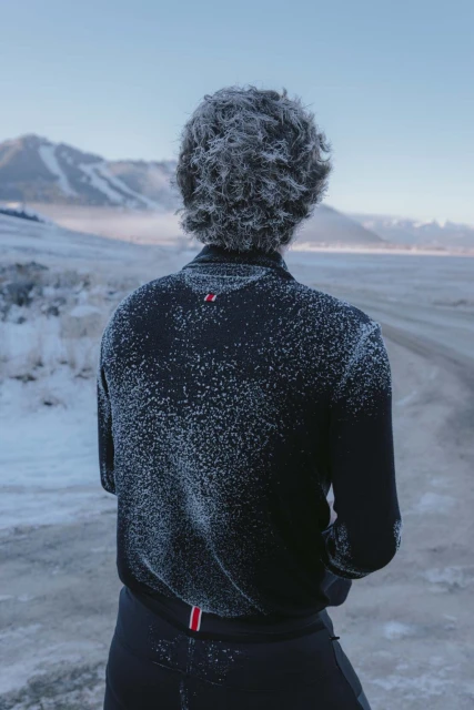 Person with frost-covered hair and jacket stands facing a snowy landscape with mountains in the distance on a clear day.