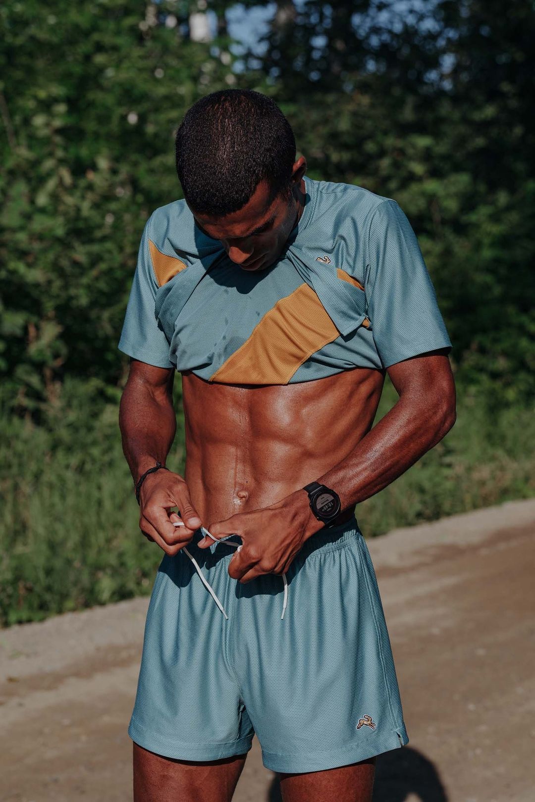 Fit, toned African American man tying shorts outside on a path with green trees and foliage in background, wearing athletic clothing.