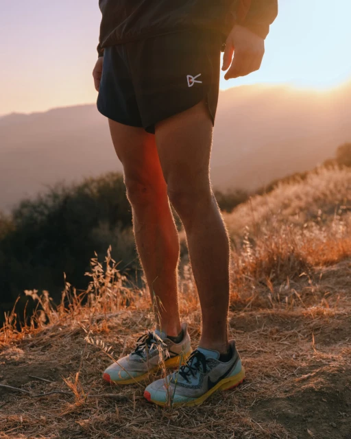 Close-up of a runner's legs and feet wearing athletic shoes and shorts, with a hilly landscape and sunset in the background.
