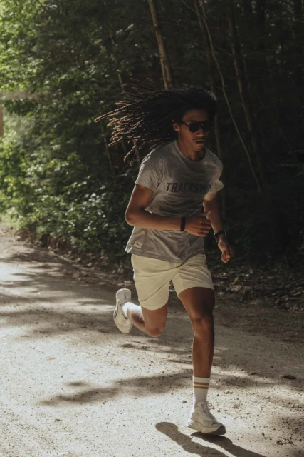 Athletic man running on dirt path in woods, wearing sunglasses, grey t-shirt, and white shorts, showcasing fitness and outdoor activity.