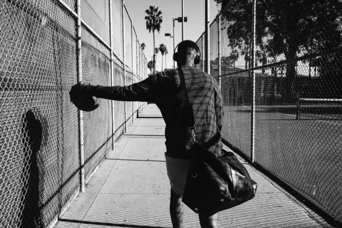 Man walking between chain-link fences holding a basketball, photographed in black and white by Emily Maye