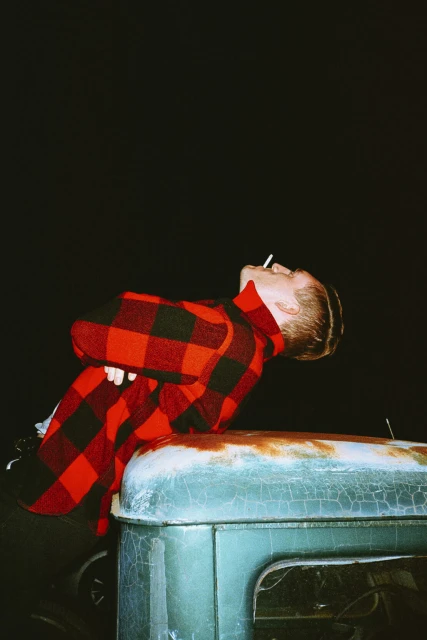 An adult male in a red and black plaid shirt leans on the hood of a weathered turquoise vehicle while smoking under a dark sky.