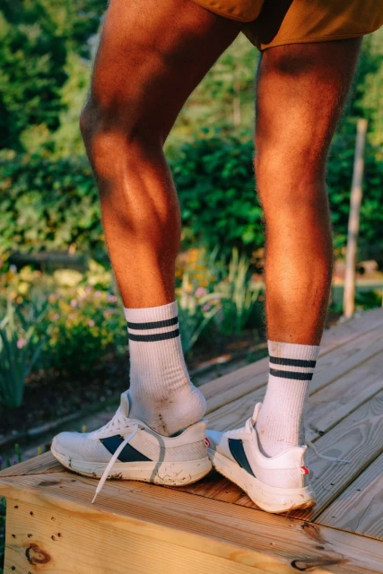 Man's legs and feet clad in white athletic shoes and striped socks standing on wooden steps outside in a green, leafy setting.