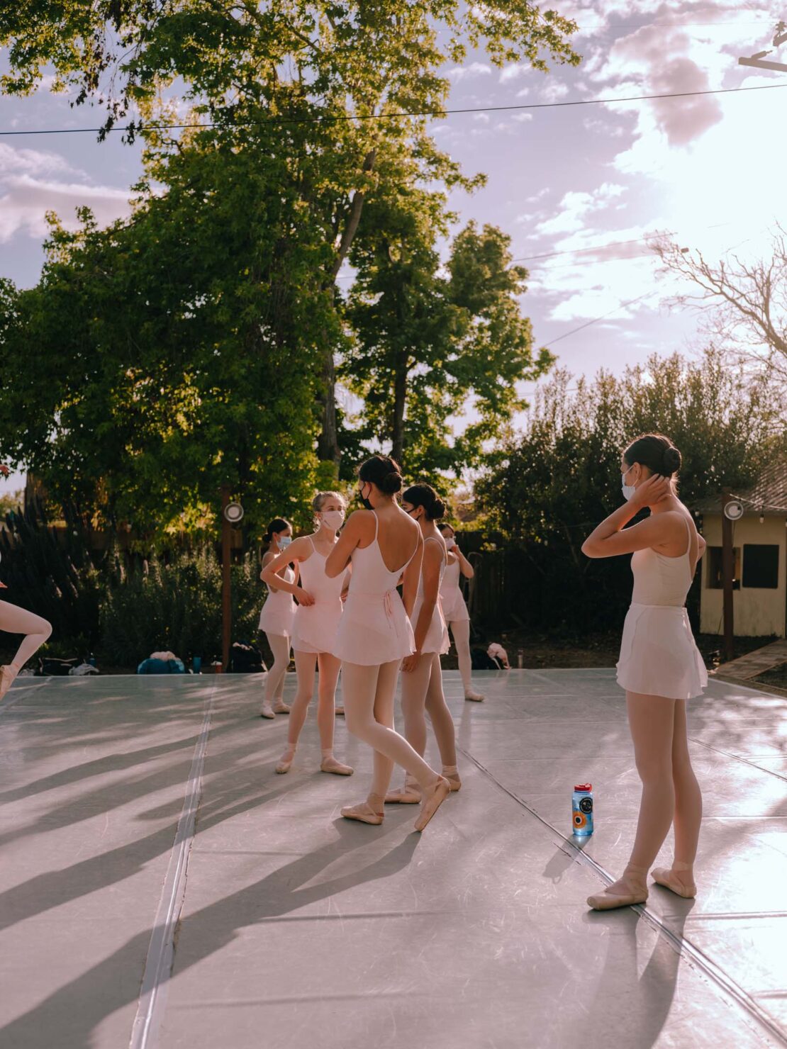 Outdoor ballet class with group of young women wearing pink dresses and face masks practicing in a grassy area under a sunny sky.