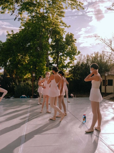 Outdoor ballet class with group of young women wearing pink dresses and face masks practicing in a grassy area under a sunny sky.