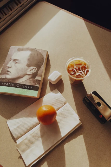 A still life of a book by Frank O'Hara, a plastic cup of iced coffee, an orange, a pair of earbuds, and a cassette player.